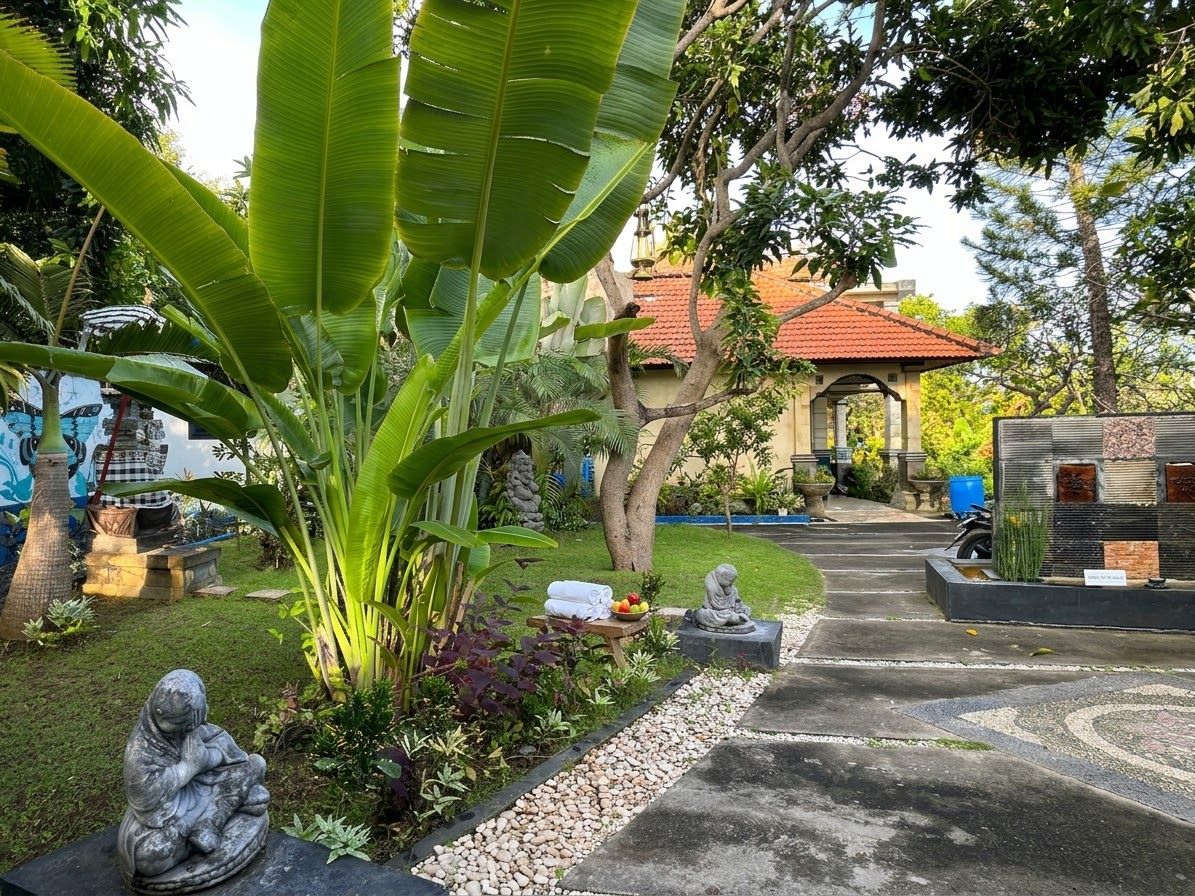 A garden pathway leading to a building with a red-tiled roof, surrounded by lush greenery and stone statues.