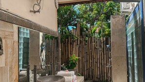 Une salle de bain avec un grand lavabo en pierre, un miroir et un espace douche délimité par une clôture en bambou.