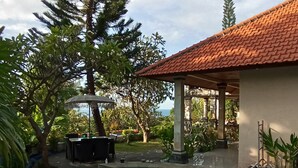 A swimming pool with a curved edge, surrounded by a garden with various plants and trees. There is a covered seating area with a table and chairs. - Charming 3-bedroom villa with WiFi, AC in the Air Sanih, The north of Bali (Bali)