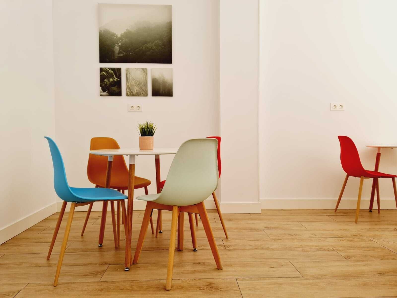 Ein Essbereich mit einem weißen Tisch, vier Stühlen in verschiedenen Farben, einer kleinen Pflanze auf dem Tisch und drei gerahmten Bildern an der Wand.