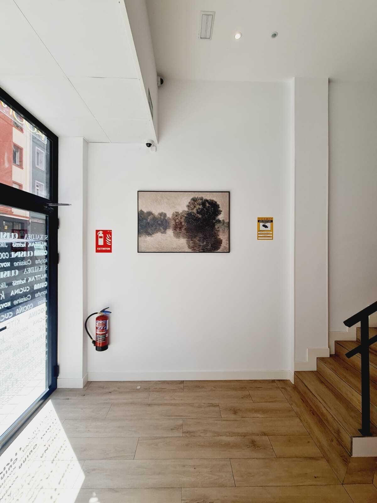 A hallway with a wooden floor, a fire extinguisher, a ceiling-mounted camera, and a framed painting on the wall.