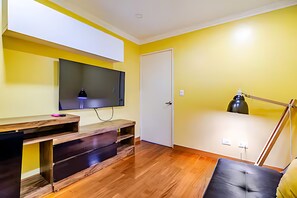 Una habitación con piso de madera, un televisor de pantalla plana montado en la pared, un escritorio con cajones, una puerta y una lámpara.