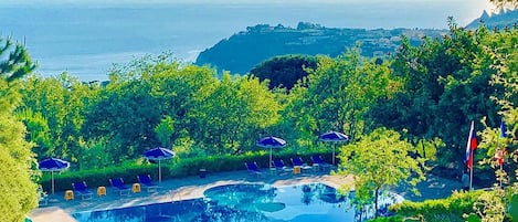 A pool area with lounge chairs and umbrellas, surrounded by greenery and overlooking a body of water.