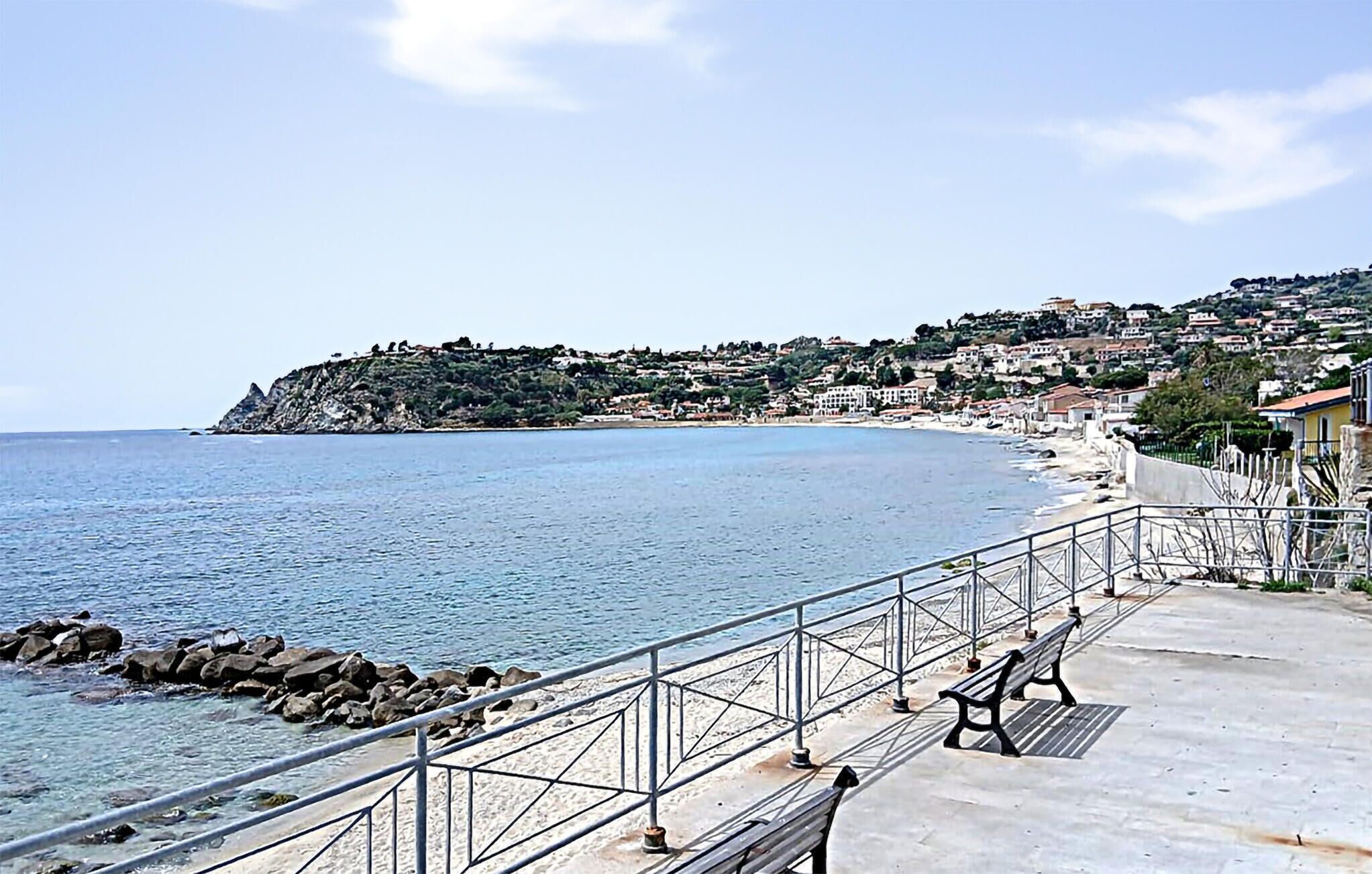 A coastal promenade with a metal railing, a bench, and a view of a hillside settlement.