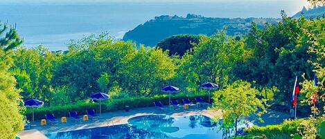 A pool area with lounge chairs and umbrellas, surrounded by greenery and overlooking a body of water.