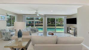 A spacious living room with a large window overlooking a pool, a ceiling fan, and a flat-screen TV mounted on the wall.