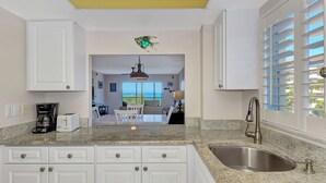 A kitchen with white cabinets, a granite countertop, a stainless steel sink, and a view of the ocean through large windows. - Playing Hooky | Beachfront condo w/ resort-style amenities, Views of the Gulf (Siesta Key)