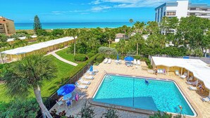 A pool area with lounge chairs and umbrellas, surrounded by greenery and a view of the ocean. - Playing Hooky | Beachfront condo w/ resort-style amenities, Views of the Gulf (Siesta Key)