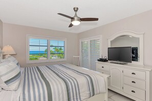 A bedroom with a bed, a ceiling fan, a TV, a dresser, and a window with a view of the ocean.