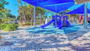 A playground with a slide and climbing structures, surrounded by trees and covered by blue shade cloths.