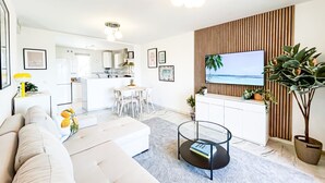 Ein modernes Wohnzimmer mit einer Couch, einem Couchtisch und einem an einer Holzwand montierten Fernseher. Im Hintergrund befindet sich eine Küche mit weißen Schränken sowie ein Essbereich mit Tisch und Stühlen.