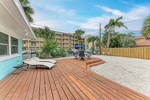 Une terrasse en bois avec des chaises longues et un espace barbecue, entourée d’une clôture en bois et de palmiers.
