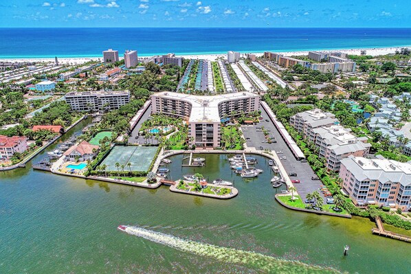 An aerial view of a coastal resort with a marina, beachfront hotels, and a central building with a unique architecture.