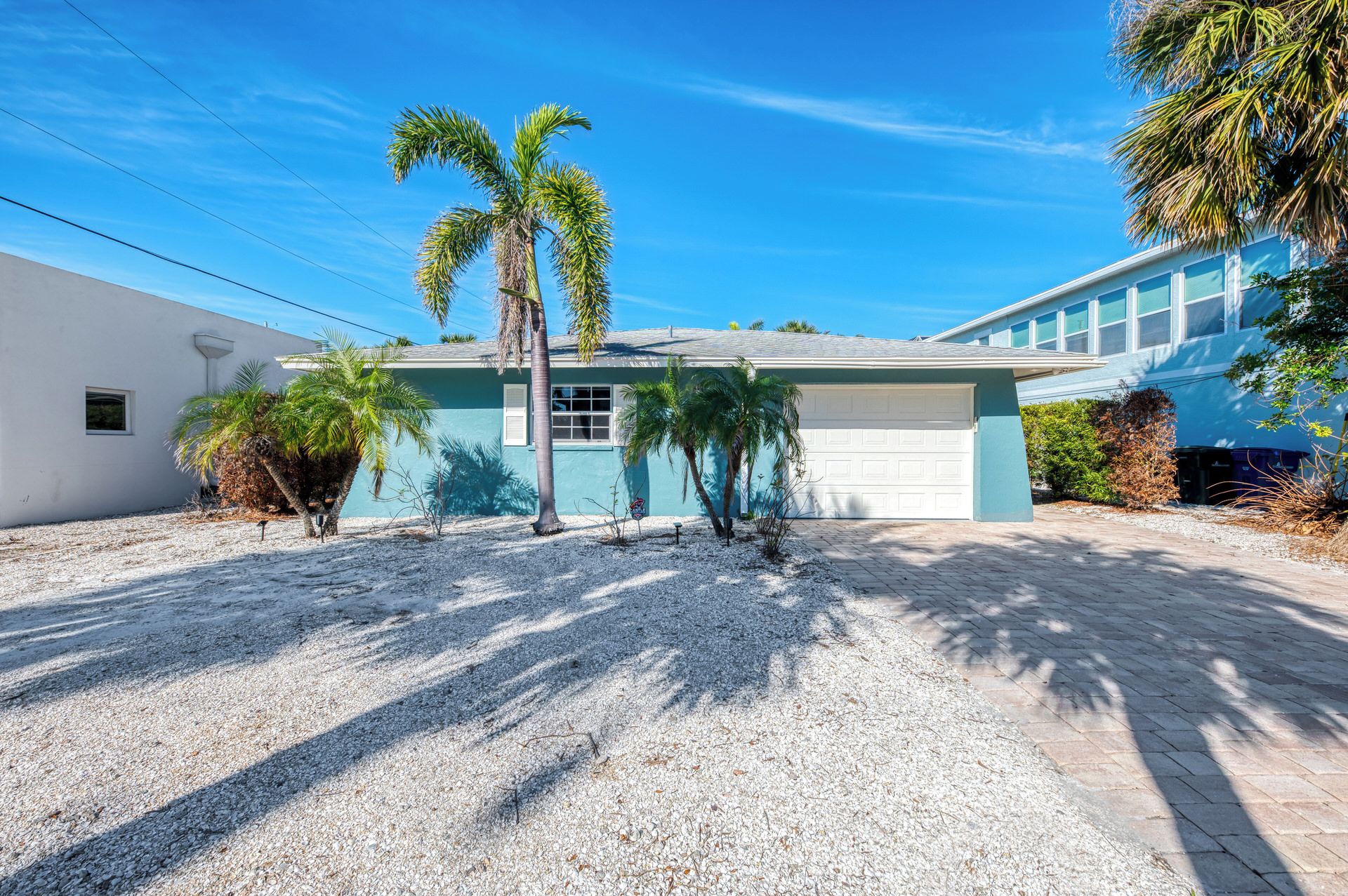 A single-story house with a garage, surrounded by gravel and palm trees.