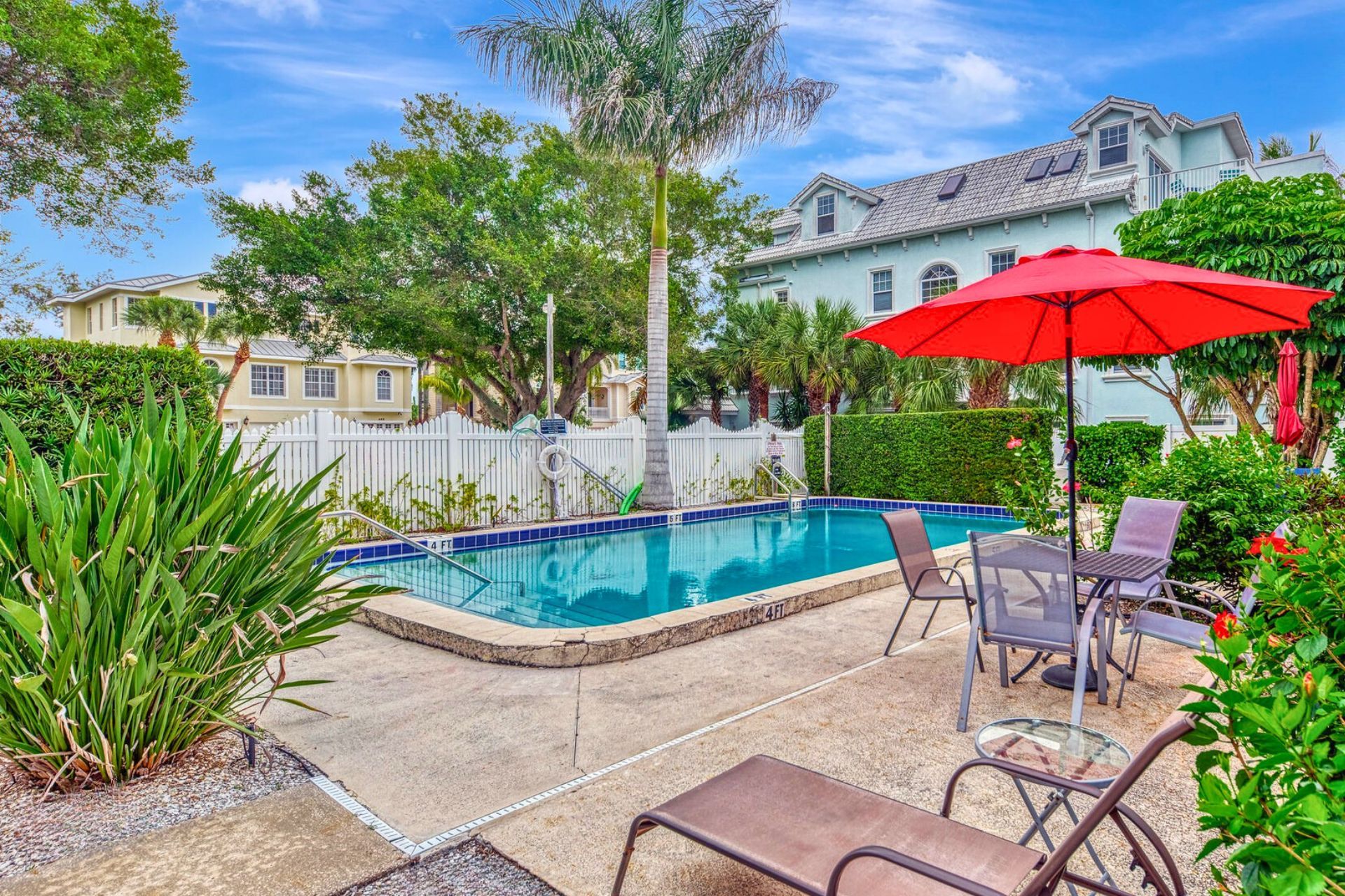A swimming pool with a 4 FT depth, surrounded by a white fence, with chairs and a red umbrella.