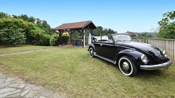 Une voiture noire d’époque est garée dans un espace verdoyant, avec un kiosque en bois et un chemin de pierre en arrière-plan.
