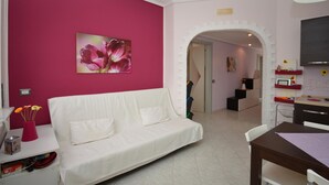 A living room with a white sofa, a pink wall, and a doorway leading to a kitchen area. - Amazing apartment in Santa Croce Camerina (Santa Croce Camerina)