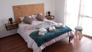 Un lit bien fait, avec une tête de lit en bois, plusieurs oreillers, un plaid turquoise et des serviettes enroulées posées sur le lit. On y trouve deux tables de chevet avec des lampes et un petit tabouret.