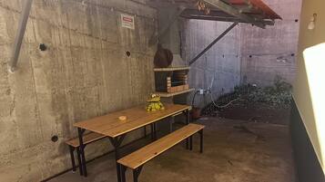 Un espace extérieur aménagé en béton, avec une table et des bancs en bois, un petit jardin avec des plantes en pot et une structure métallique au-dessus.