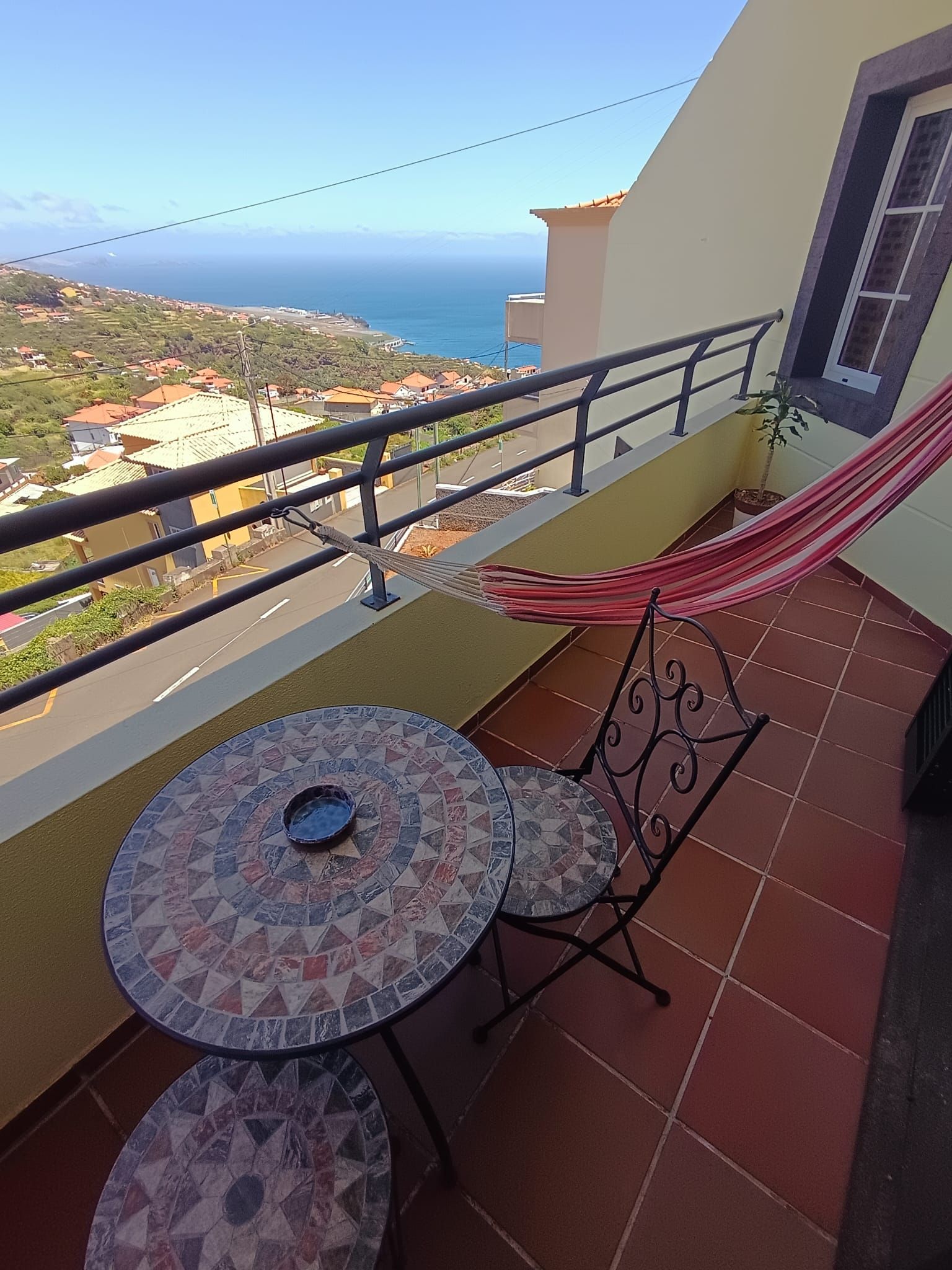 A balcony with a view of the sea, a hammock, and two mosaic-tiled tables.