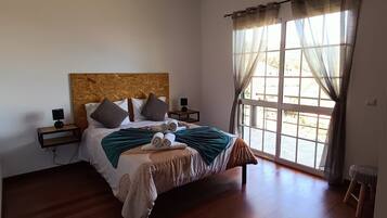 A bedroom with a wooden bed, a nightstand, a lamp, a window with curtains, and a hanging light fixture.