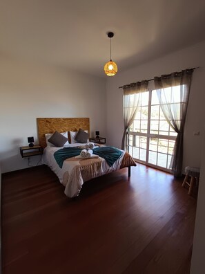 A bedroom with a wooden bed, a nightstand, a lamp, a window with curtains, and a hanging light fixture. - Dotty's House (Santa cruz)