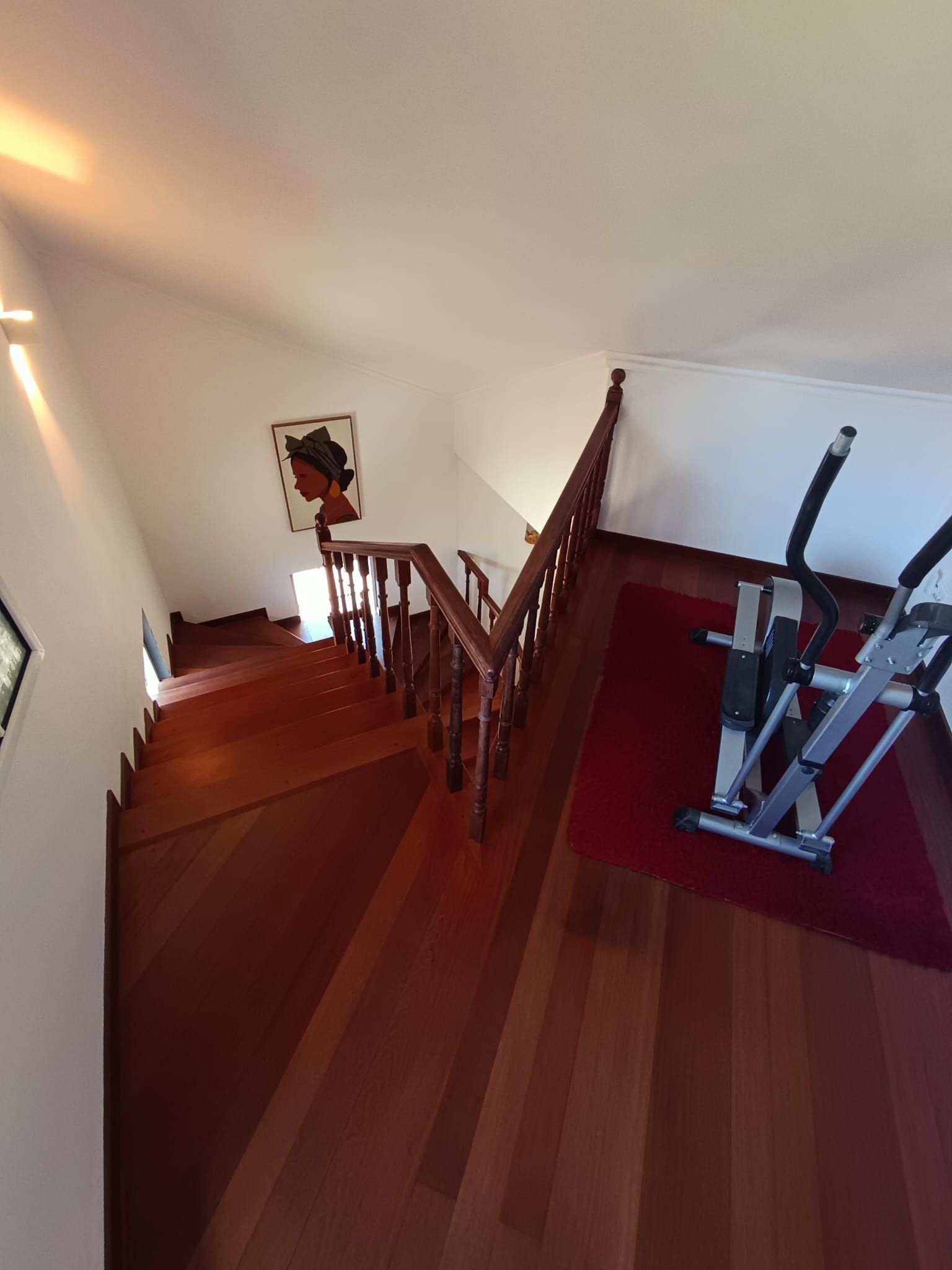 Een trap met houten leuningen die uitkomt op een plateau, waar een loopband op een rode mat staat.