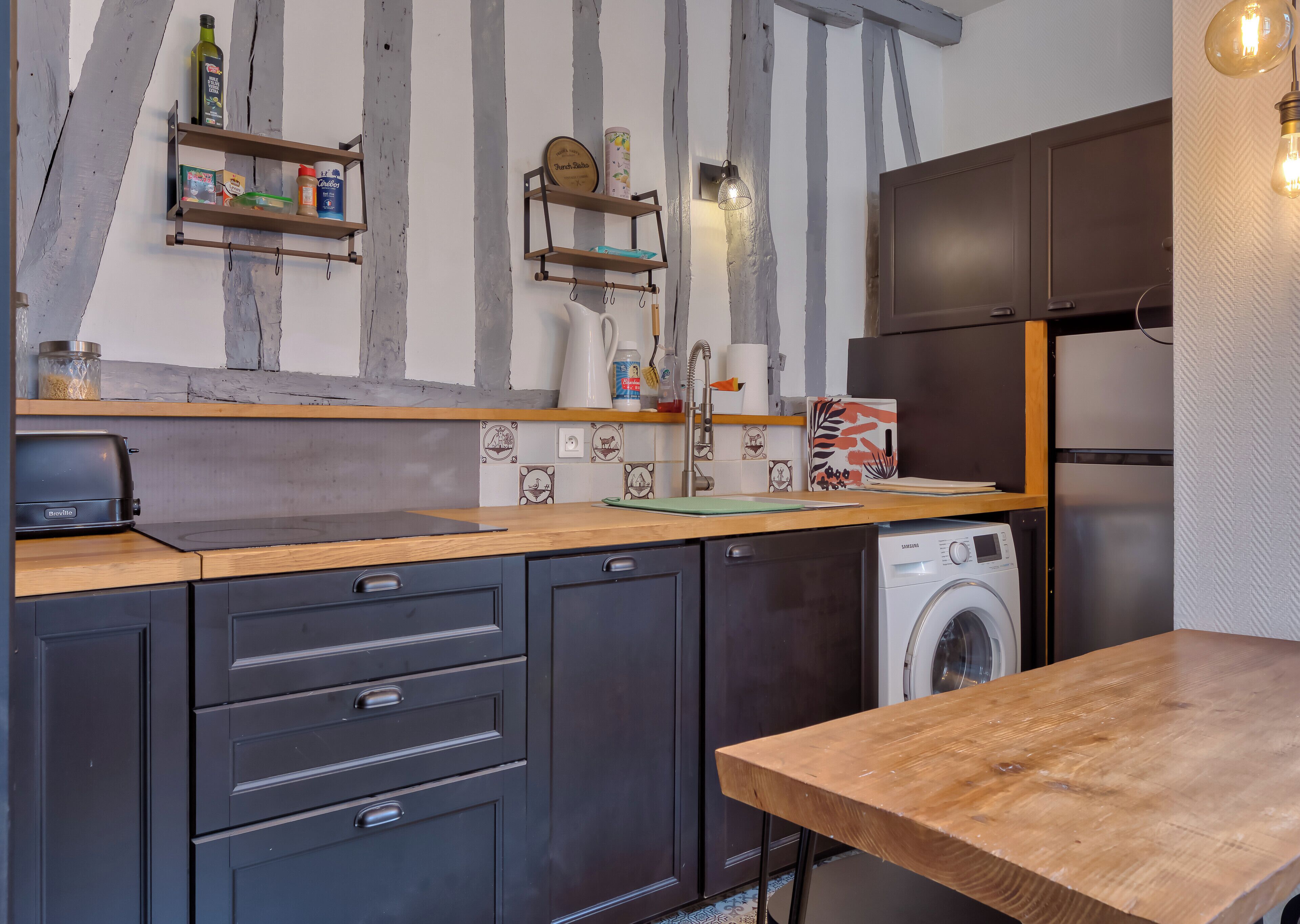 A modern kitchen with dark cabinets, a wooden countertop, a Breville toaster, a Samsung washing machine, and a wooden dining table.