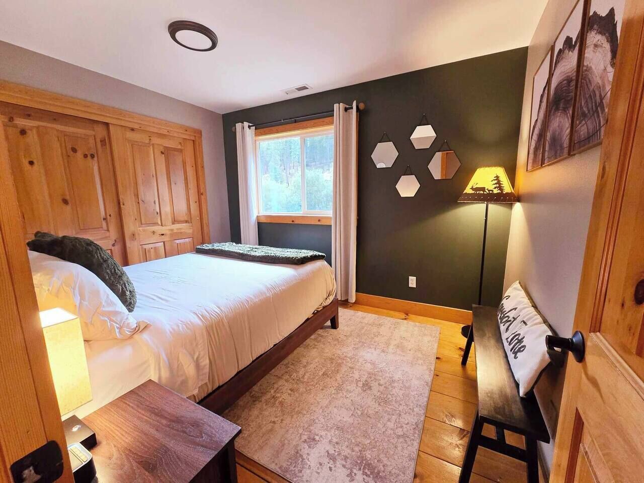 A bedroom with a bed, wooden wardrobe, a bench, a lamp, and a window with curtains.