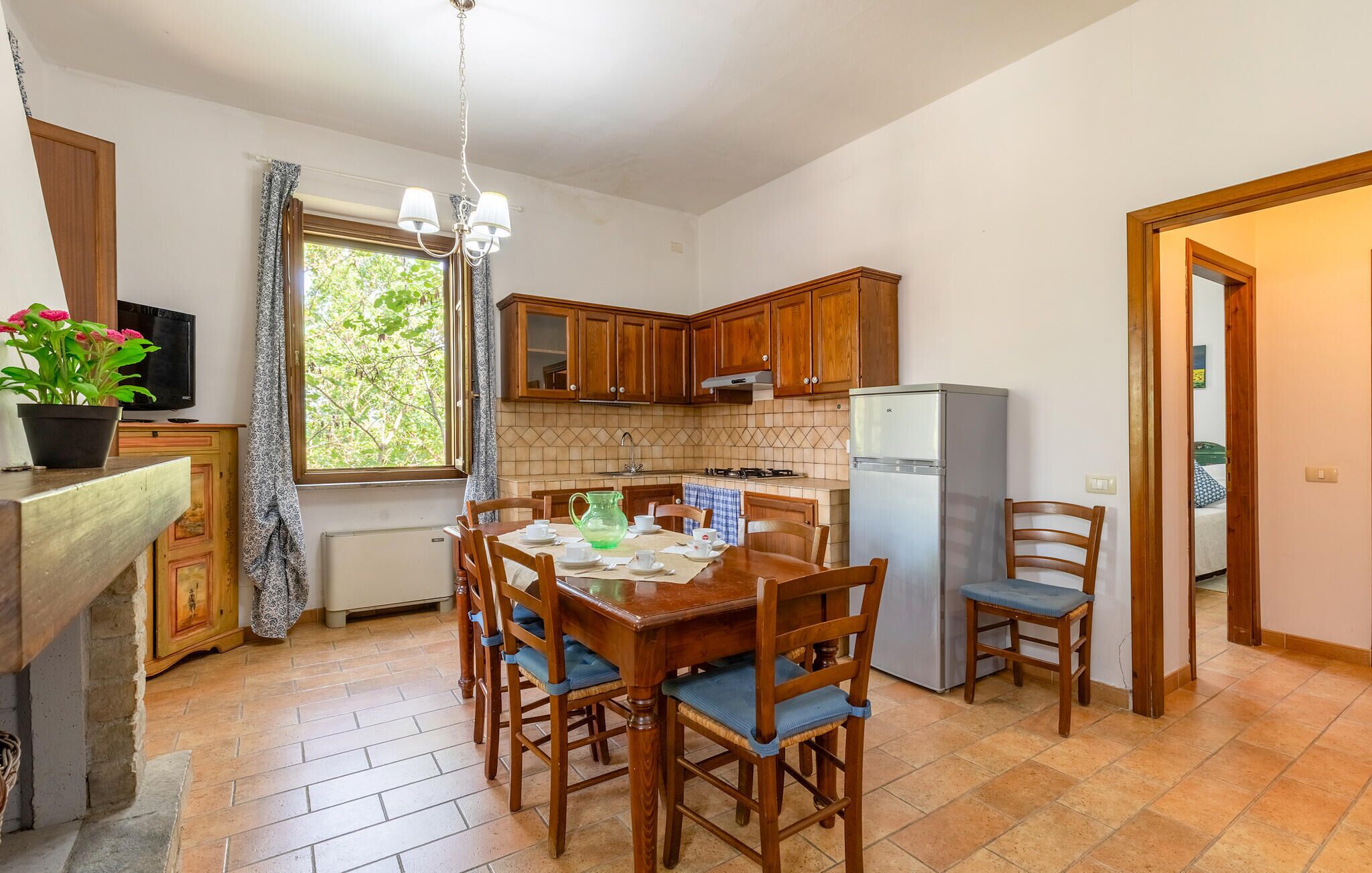 Une cuisine avec des meubles de rangement en bois, une table à manger avec des chaises, un réfrigérateur et une fenêtre avec des rideaux.