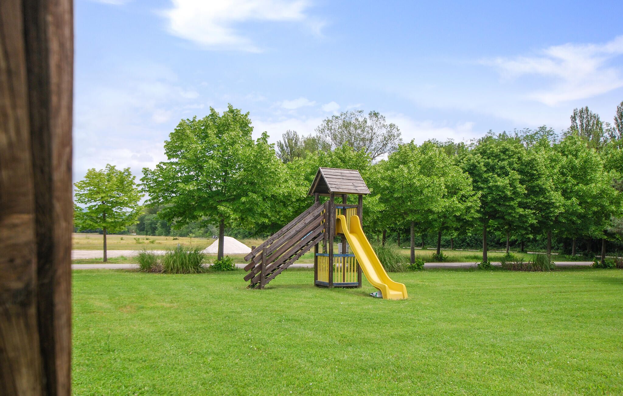 Une aire de jeux avec un toboggan en bois et une structure d’escalade, entourée d’herbe verte et d’arbres.