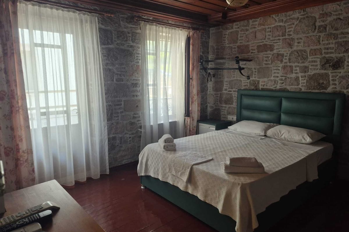 A bedroom with a stone wall, a wooden ceiling, a green upholstered headboard, a bed with white linens, and a wooden nightstand with a lamp.