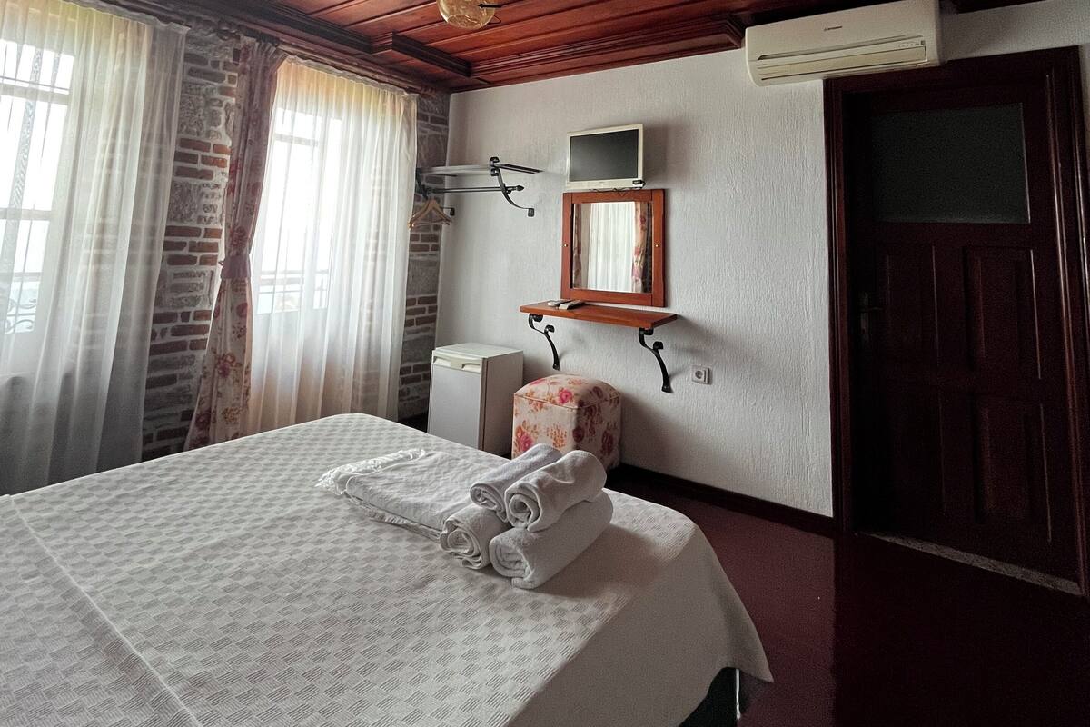 A bedroom with a wooden ceiling, a bed with white linens, a small bedside table, a mirror, and a window with curtains.