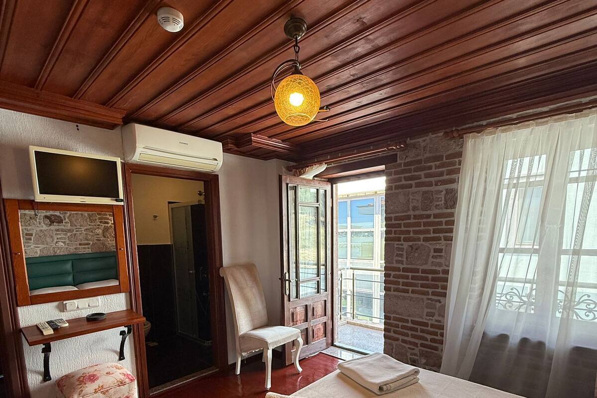 A bedroom with a wooden ceiling, a bed with a white bedspread, a chair, a small table with a remote control, and a stone wall.