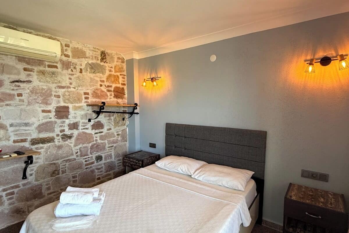 A bedroom with a stone wall, a bed with white linens, and a nightstand with a lamp.