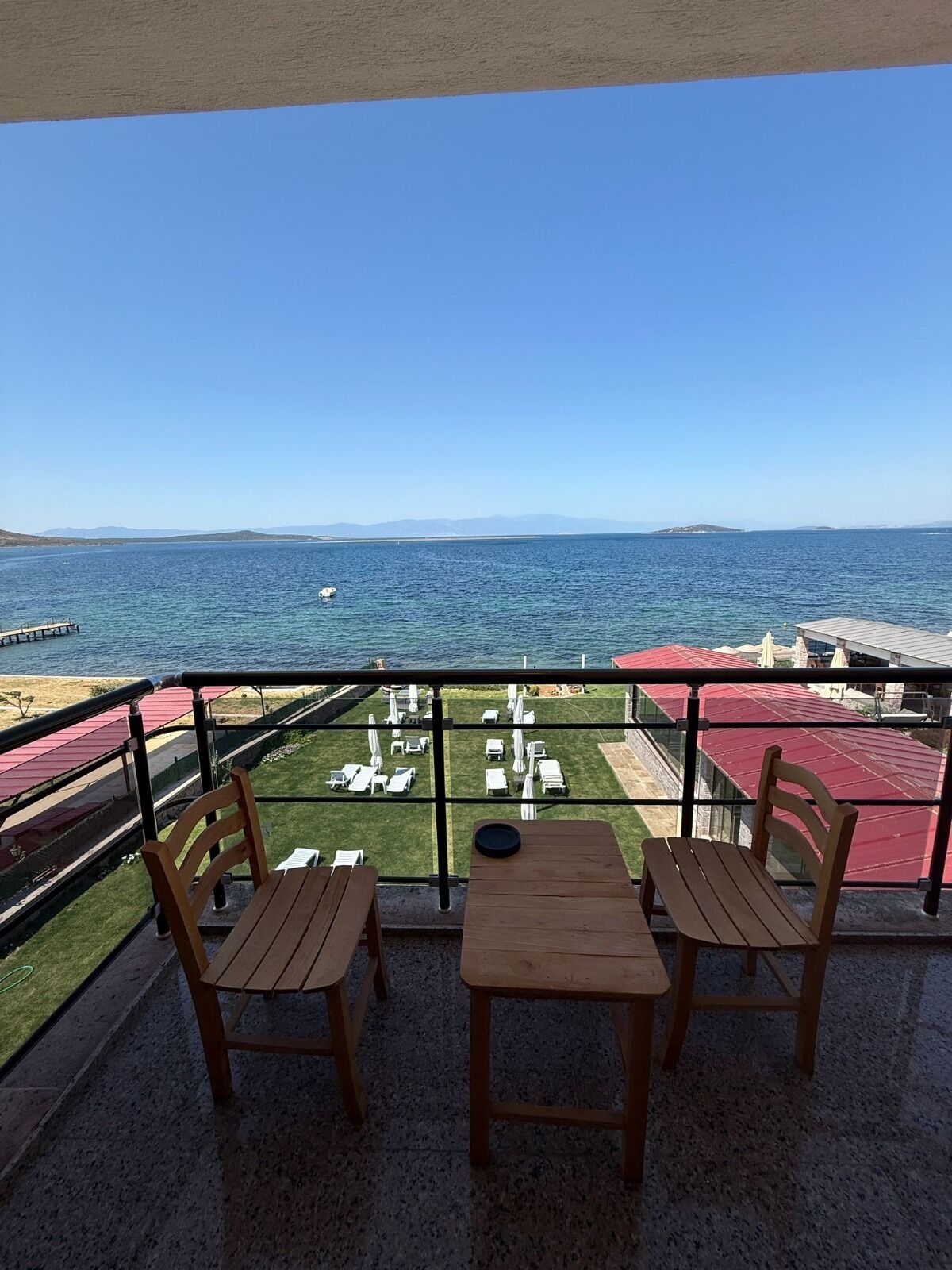 A balcony with two wooden tables and chairs, overlooking a beach with a clear view of the sea and distant hills.
