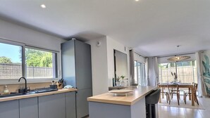 Eine moderne Küche mit einer zentralen Kochinsel, einem Essbereich mit einem Holztisch und Stühlen, sowie großen Fenstern, die für natürliches Licht sorgen.