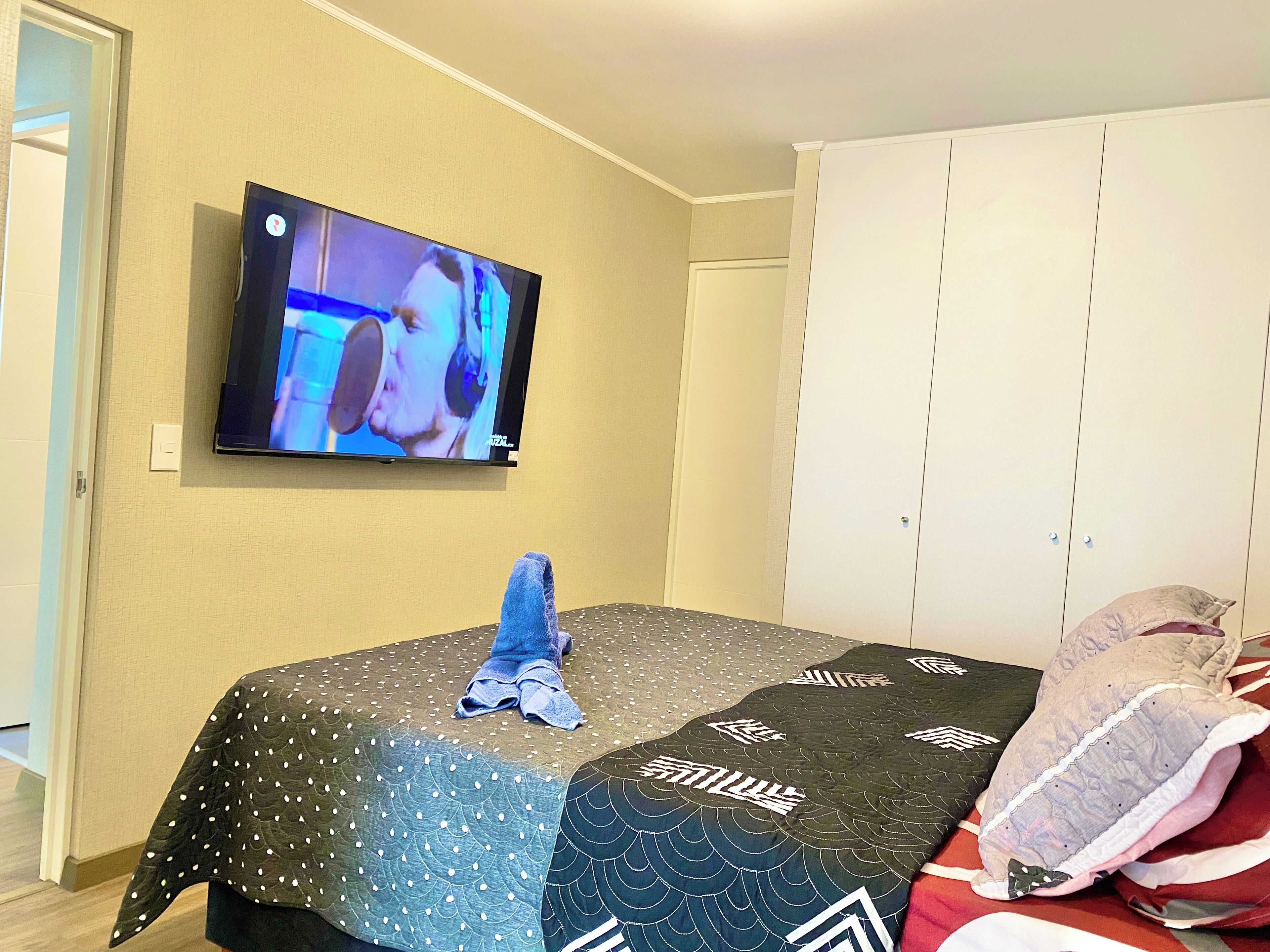 Un dormitorio con una cama cubierta por un colcha, un televisor en la pared y un armario.