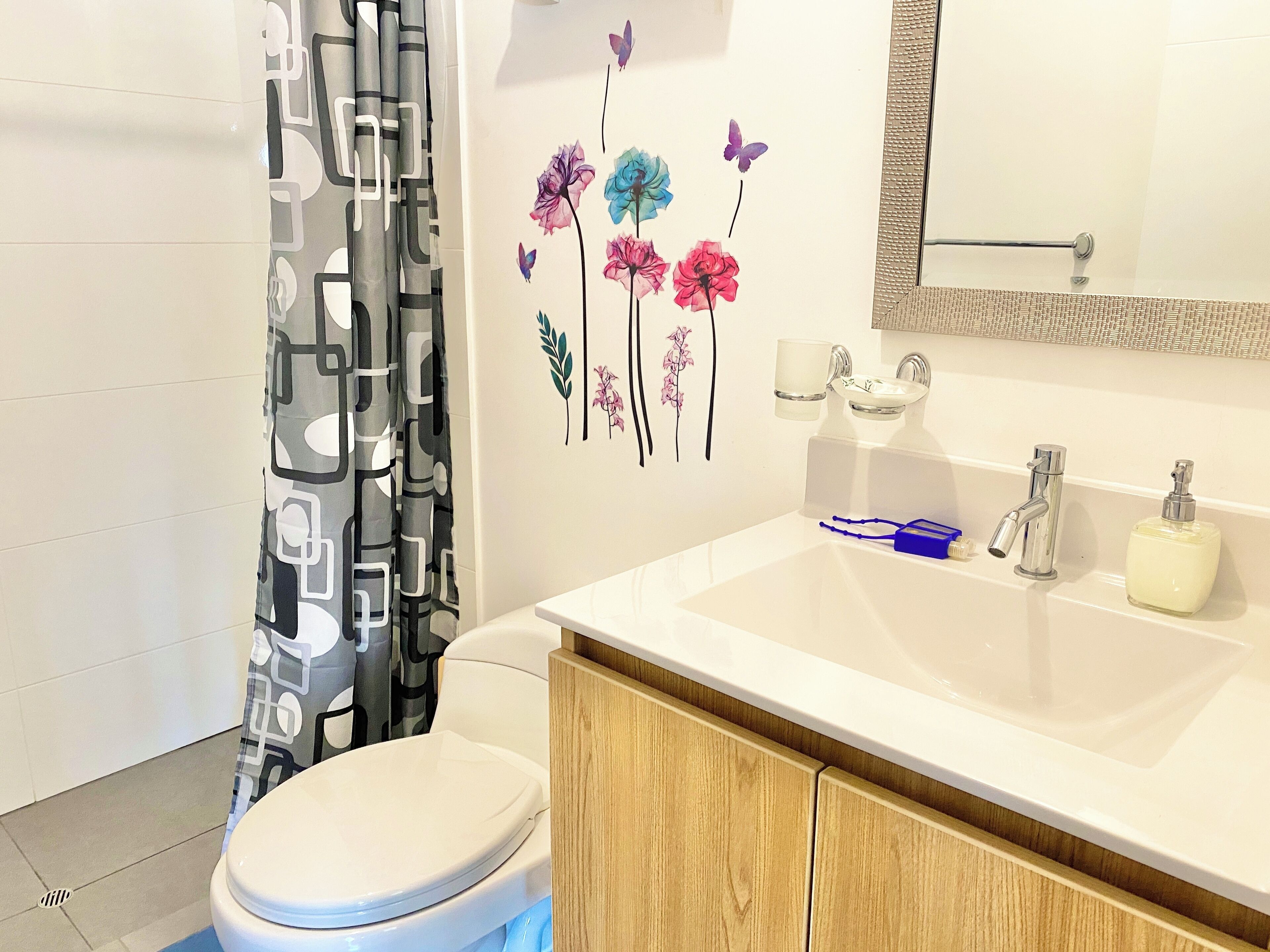 Une salle de bain avec des toilettes blanches, un meuble en bois, un lavabo blanc avec un miroir au-dessus, et un rideau de douche à motifs géométriques.