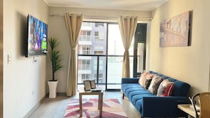 A living room with a TV, a sofa, a table, a potted plant, and a view of buildings through a window.