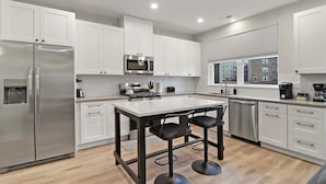 A modern kitchen with white cabinets, stainless steel appliances, a central island with stools, and a view of the city through the window. - Warrenville Rep 2 (Warrenville)