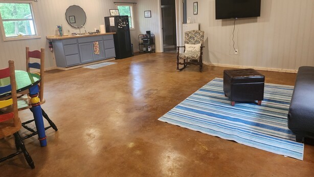 A spacious living room with a wooden floor, a kitchen area with a grey cabinet, a black refrigerator, a blue and white striped rug, and a black leather ottoman. - 1-bedroom, dog friendly cottage (Hot Springs)