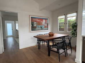 A dining area with a wooden table and chairs, a framed poster on the wall, and a view through the window.