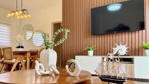 A modern dining area with a round wooden table, white decorative items, a television mounted on a wooden wall, and a potted plant.