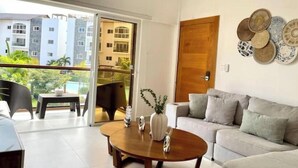 A modern living room with a sofa, a coffee table, and a dining area with chairs. There is a sliding glass door leading to a balcony with a view of buildings and greenery.