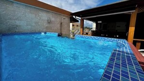 Una piscina en la azotea con agua azul cristalina, bordeado de azulejos y con una zona de estar techada.