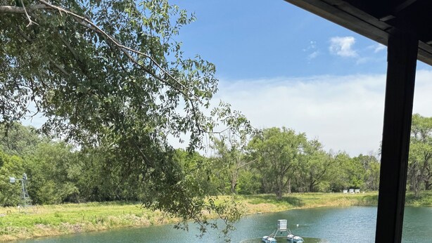 Blick von einer überdachten Außenbereich mit Tisch und zwei Stühlen auf einen ruhigen Fluss mit Booten und viel Grün.