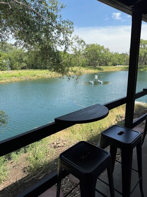 Vista desde un espacio al aire libre techado, con una mesa y dos sillas, que da a un río tranquilo con barcos y vegetación.