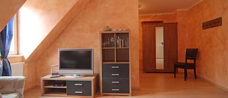 A room with a sloped ceiling, a television on a wooden stand, a bookshelf, a chair, and a door leading to another room.