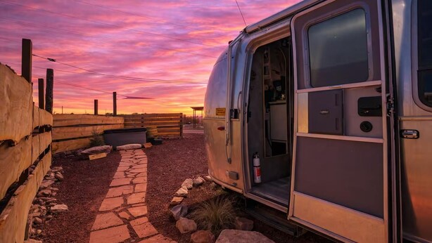 A silver RV parked in a rural area with a stone pathway leading to it, surrounded by a wooden fence and a wooden bench.